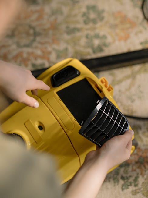 Close-up of a person's hands operating a yellow carpet cleaning machine in a room with a patterned, traditional-style rug featuring shades of beige, blue, and red. The machine appears to be a deep cle